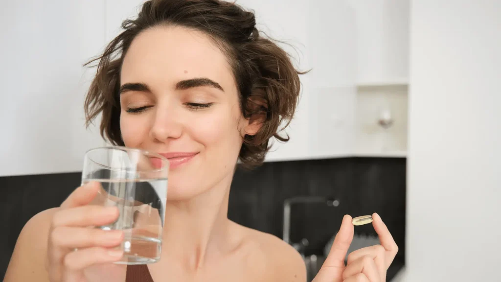 Mulher sorridente segurando um copo de água em uma mão e um comprimido na outra, prestes a tomar o remédio.