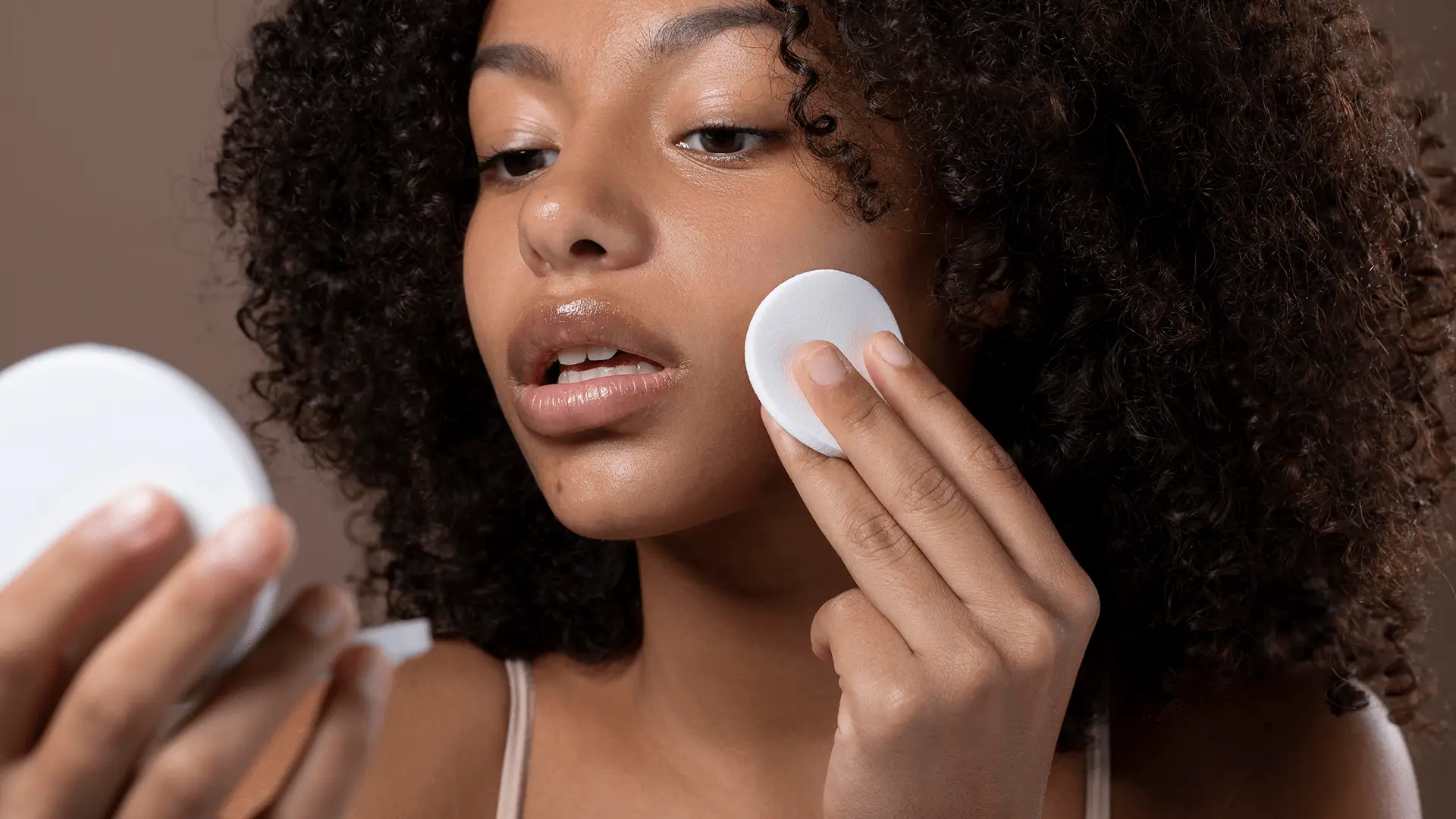 Mulher com cabelo cacheado aplica uma base para no rosto para o tom de pele usando um algodão, olhando para um espelho compacto.