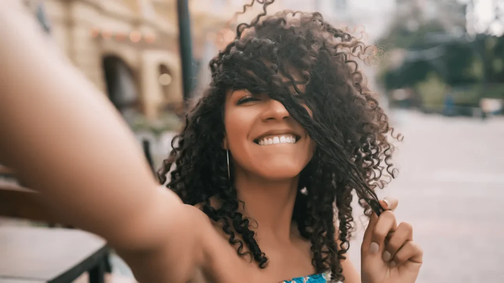 Mulher sorridente com cabelo cacheado, fazendo selfie ao ar livre, segurando um cacho de cabelo.
