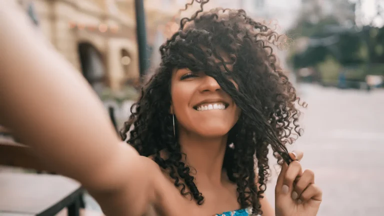 Mulher sorridente com cabelo cacheado, fazendo selfie ao ar livre, segurando um cacho de cabelo.