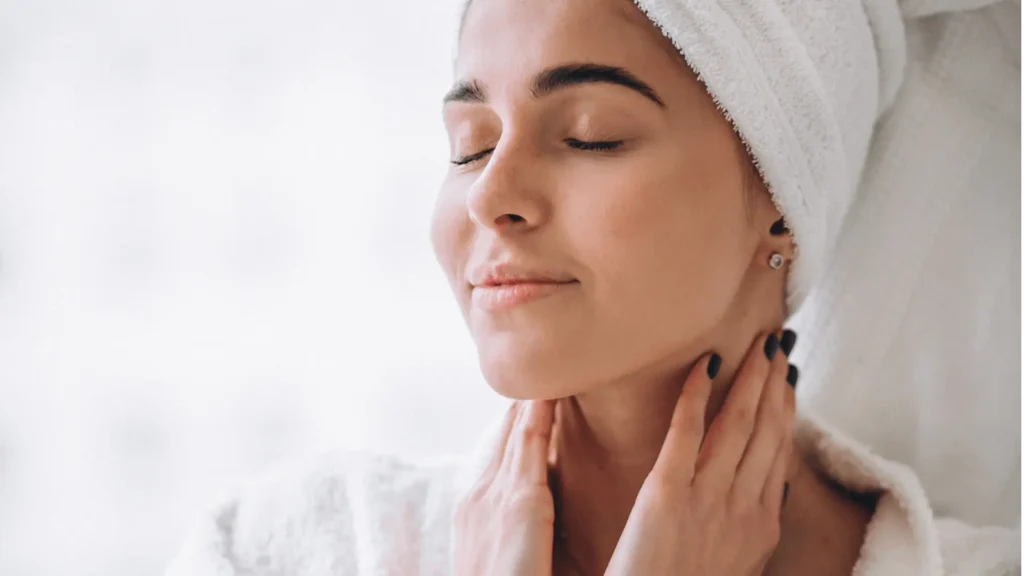 Mulher com toalha na cabeça e roupão branco, com os olhos fechados e expressão serena, massageando o pescoço e a pele.