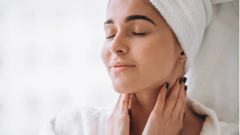Mulher com toalha na cabeça e roupão branco, com os olhos fechados e expressão serena, massageando o pescoço e a pele.