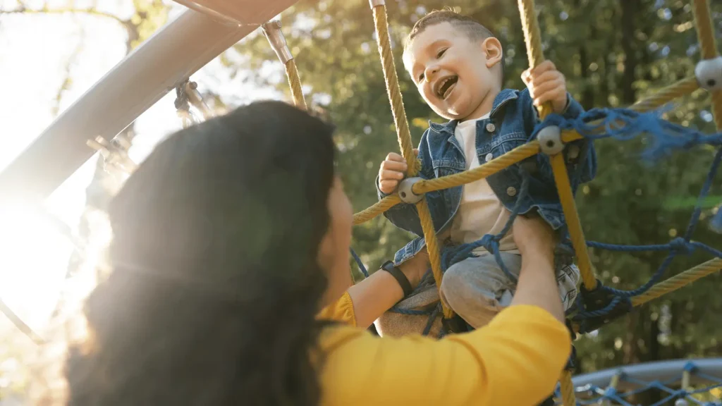 Adulto com blusa amarela ajuda criança sorridente em parque de cordas ao ar livre, sob luz solar.