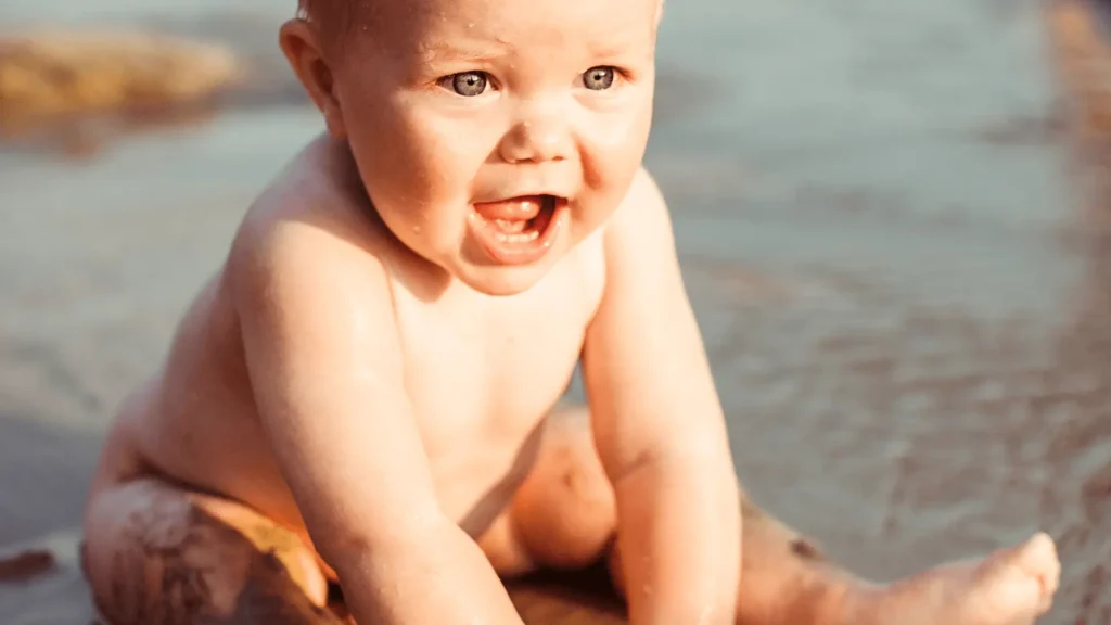 Bebê sorridente, sem roupa, sentado na areia molhada de uma praia ou lago, com água ao fundo.