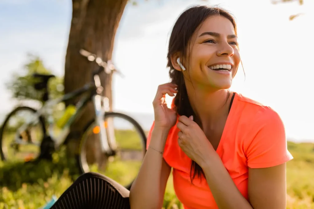 mulher jovem e sorridente, com cabelos castanhos escuros, usando uma camiseta laranja vibrante e fones de ouvido brancos. Ela parece estar sentada em um campo gramado, com uma bicicleta visivelmente em segundo plano, encostada em uma árvore.