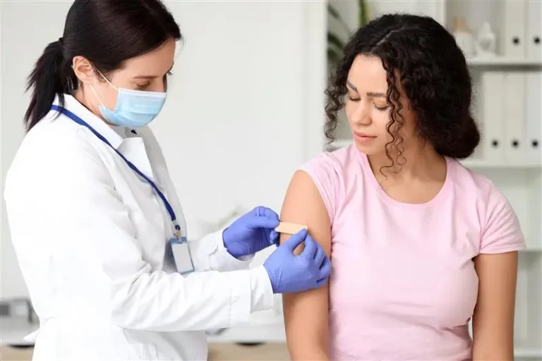 Profissional de saúde aplicando uma dose de vacina no braço de uma mulher sentada, em ambiente clínico e demonstrando a importância da vacinação.
