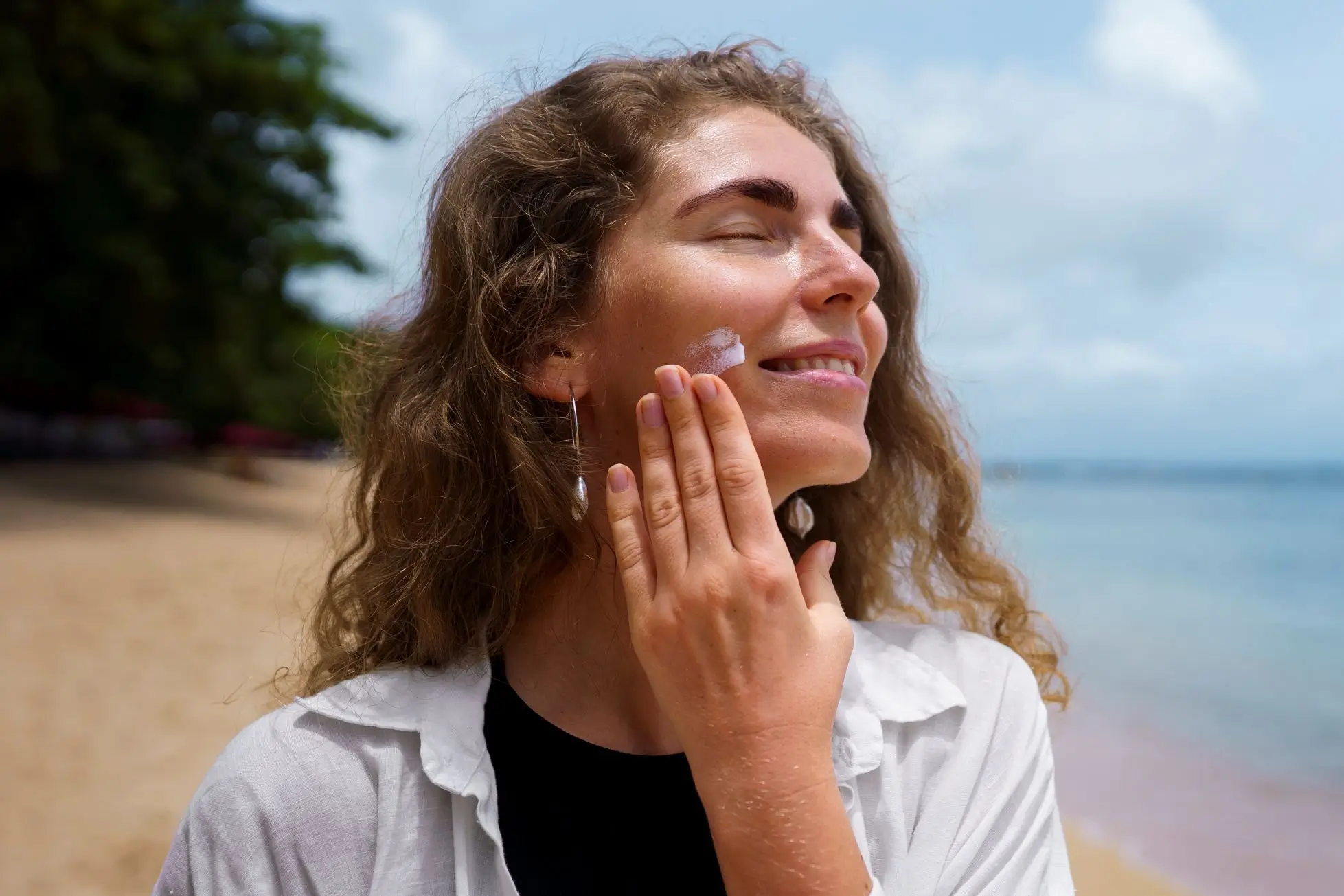 Mulher sorridente com cabelos cacheados e castanhos, aplicando protetor solar no rosto com a mão direita. Ela está na praia, com o oceano e o céu azul ao fundo. Seus olhos estão fechados e há um sorriso suave em seu rosto. Ela usa uma camisa branca de botões sobre uma blusa preta e brincos longos e prateados.
