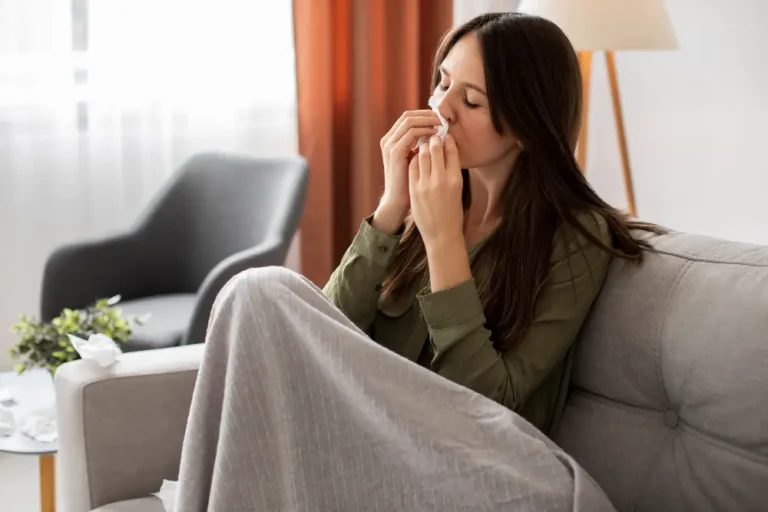 Mulher sentada em um sofá assoando o nariz com um lenço durante um quadro de congestão nasal e desconforto respiratório.
